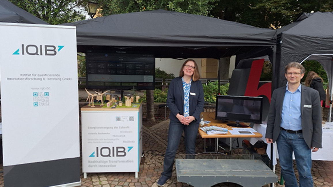 Zwei Personen stehen vor einem Messestand mit schwarzem Pavillon, Tischen, Monitor und Informationsmaterial.